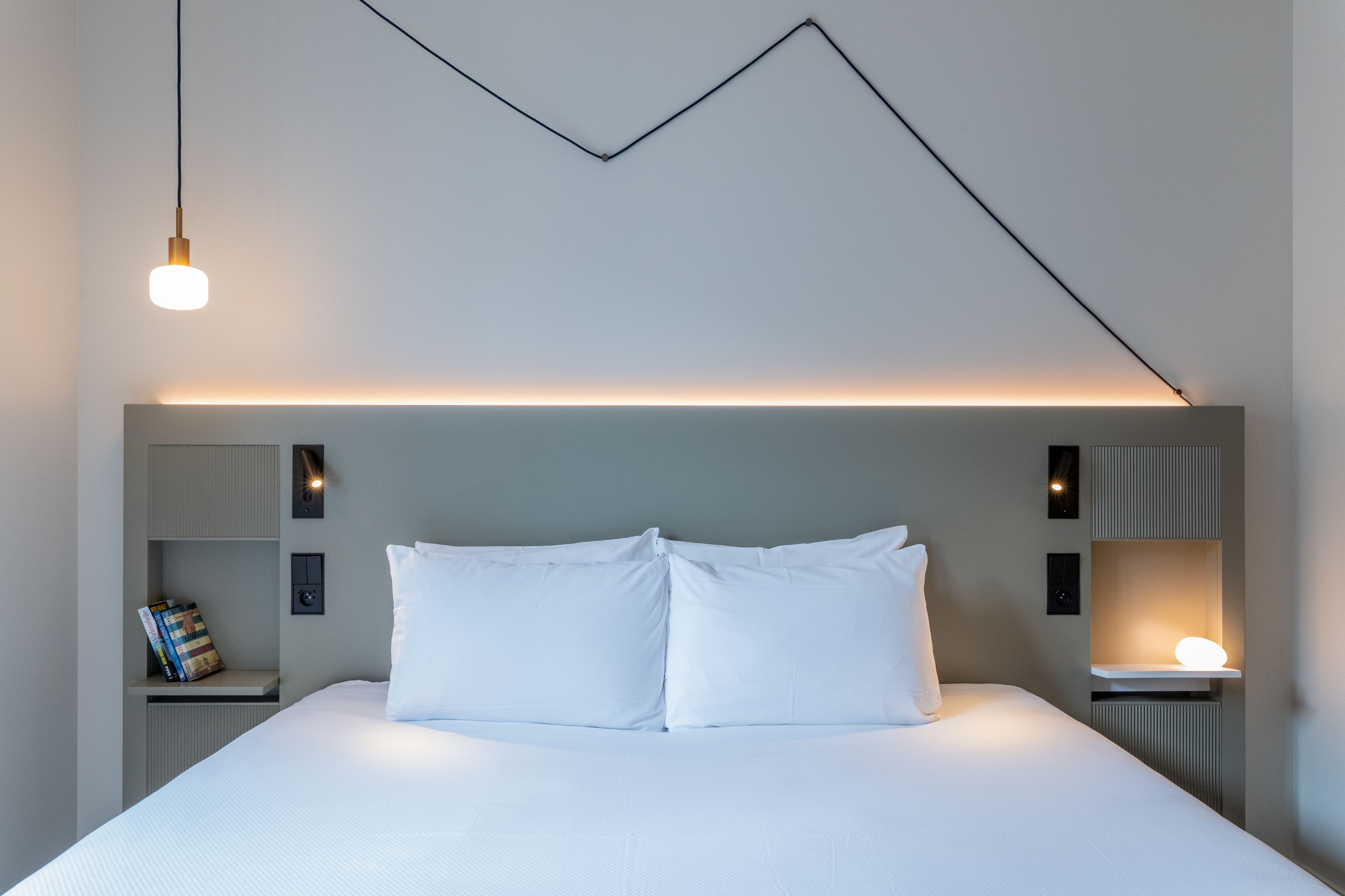 A modern hotel room with a neatly made bed, a wall-mounted light fixture, and a bedside table with a lamp.