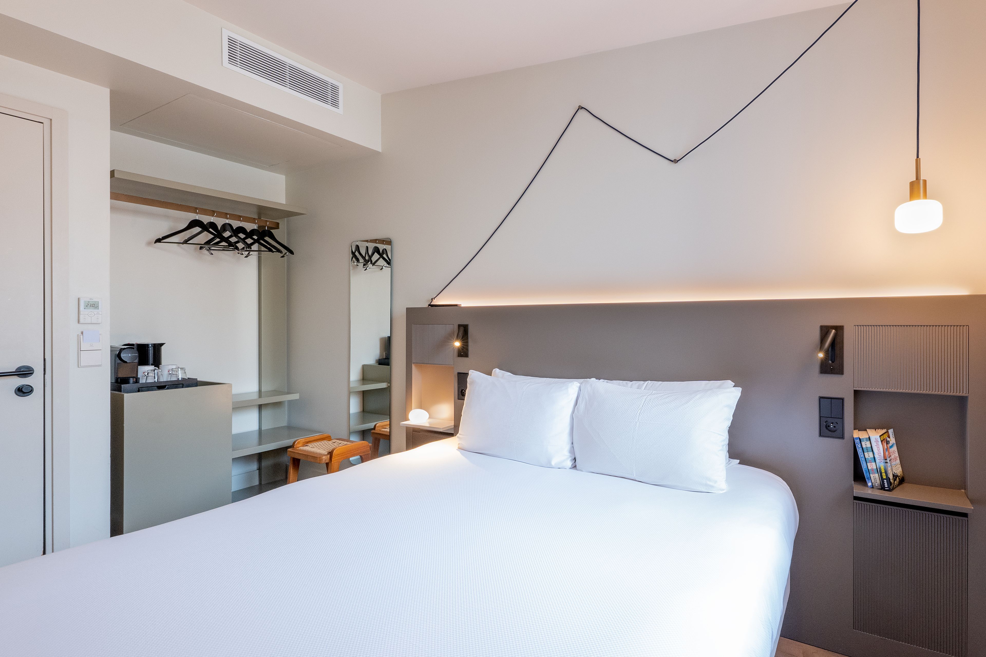 Habitación de hotel con cama, mesitas de noche, un espejo, perchero y estante en la pared con libros.
