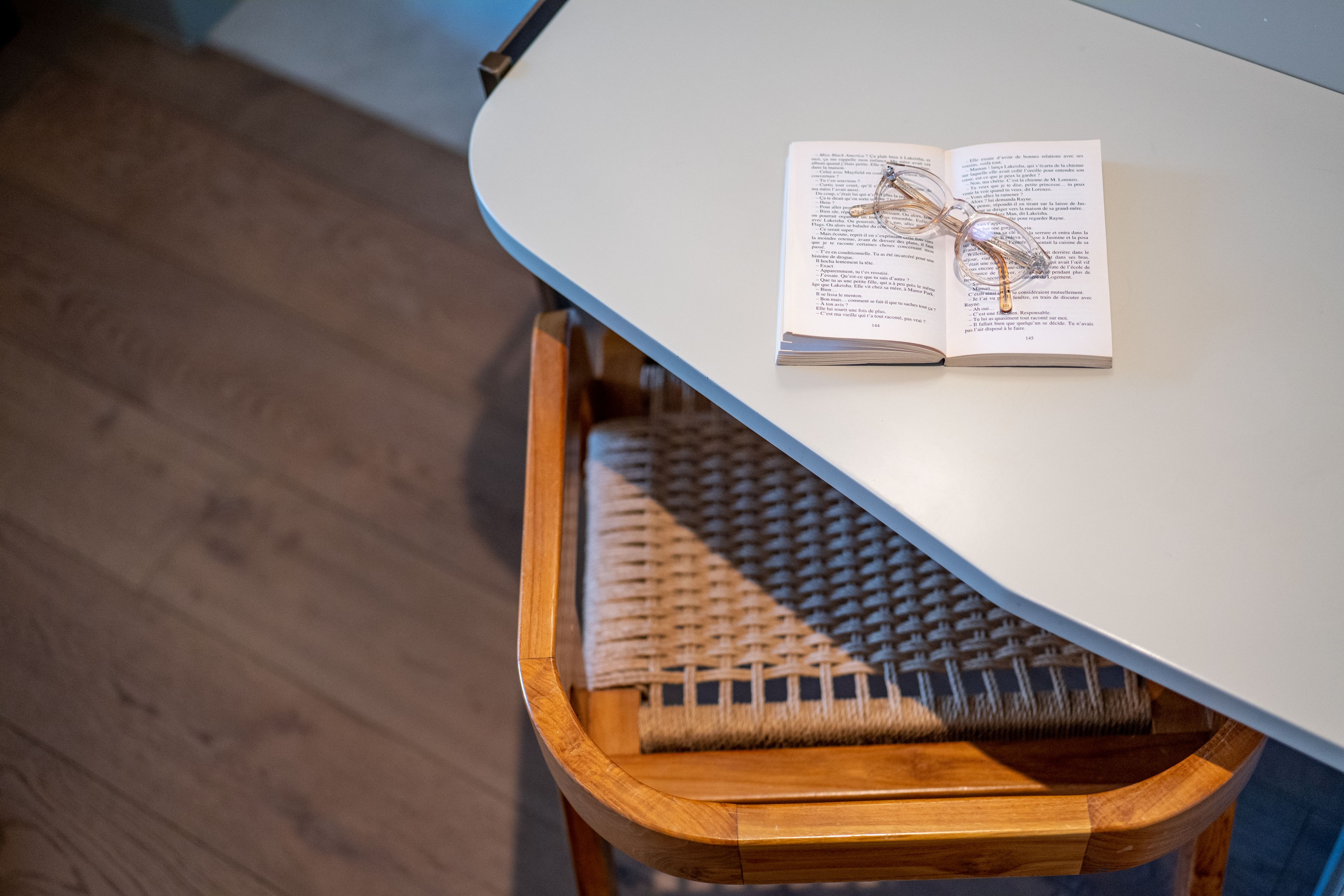 A wooden stool with a woven seat under a table, with an open book on the table.