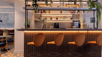 A bar area with a wooden counter, three bar stools, shelves with bottles and glasses, and hanging plants.
