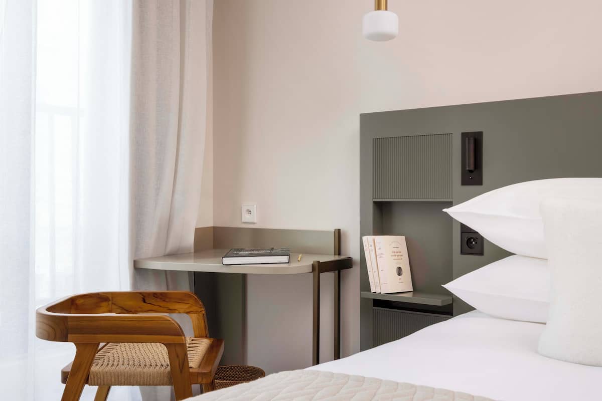 Ein ordentlich eingerichtetes Hotelzimmer mit einem Bett, einem kleinen Schreibtisch mit Stuhl, einer Lampe und einem Bücherregal.