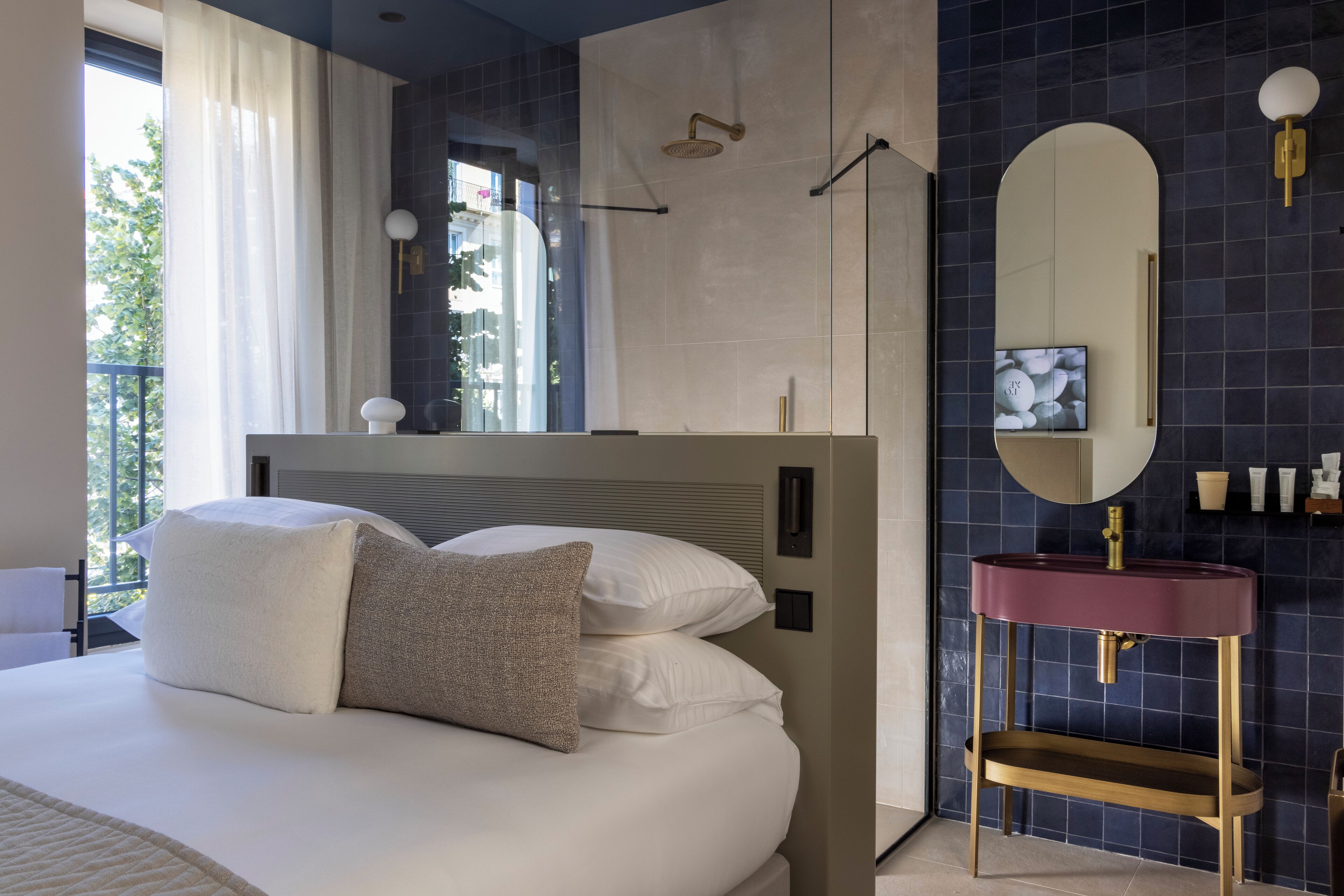 A modern bedroom with a large bed, a vanity with a mirror, and a view of greenery outside.