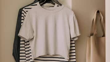Una habitación ordenada con un perchero que muestra una camiseta blanca cropped y una camisa a rayas, un bolso tote beige y una máquina de café con una taza y una dona sobre la encimera.