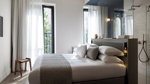 Un dormitorio moderno con una cama grande, un taburete de madera, una mesita de noche y un baño con ducha.