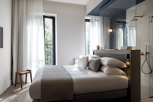 Quarto moderno com uma cama grande, um banquinho de madeira, uma mesinha de cabeceira e um banheiro com chuveiro.