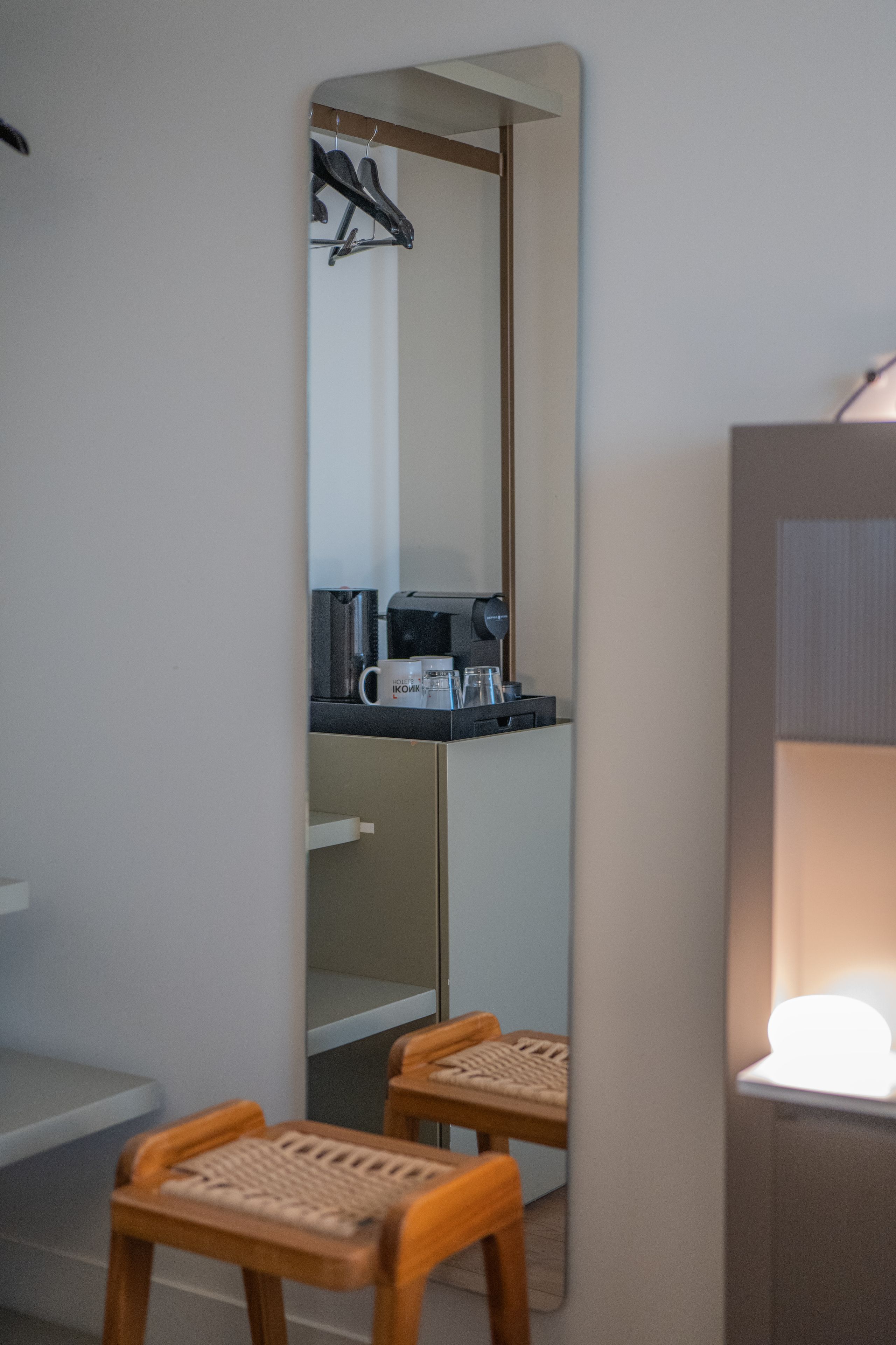 A room with a mirror reflecting a kitchenette area, a coffee machine, and a hanging coat rack. There are two wooden chairs in front of a small table.