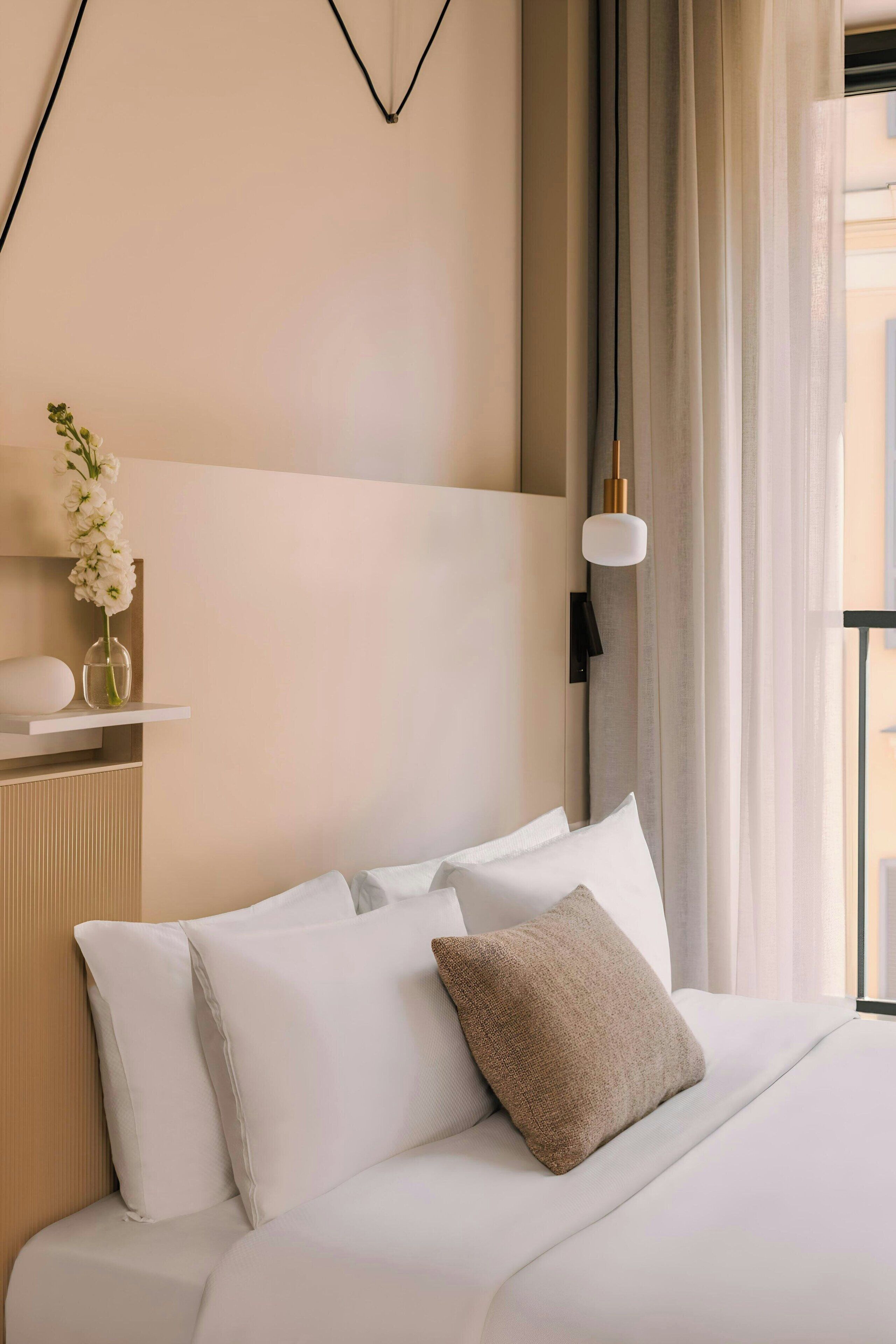 Een verzorgde hotelkamer met een bed, kussens, een vaas met bloemen en een wandlamp.