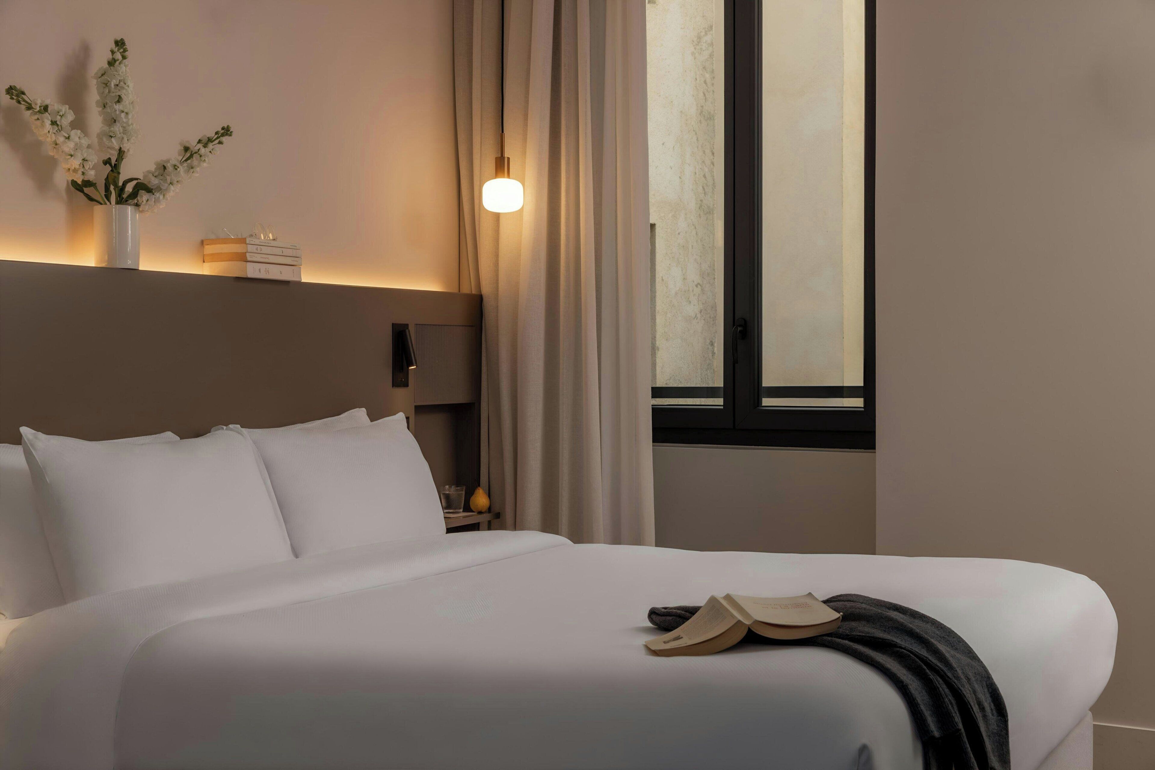 A neatly made bed with white linens, a bedside table with a lamp, and a window with curtains.