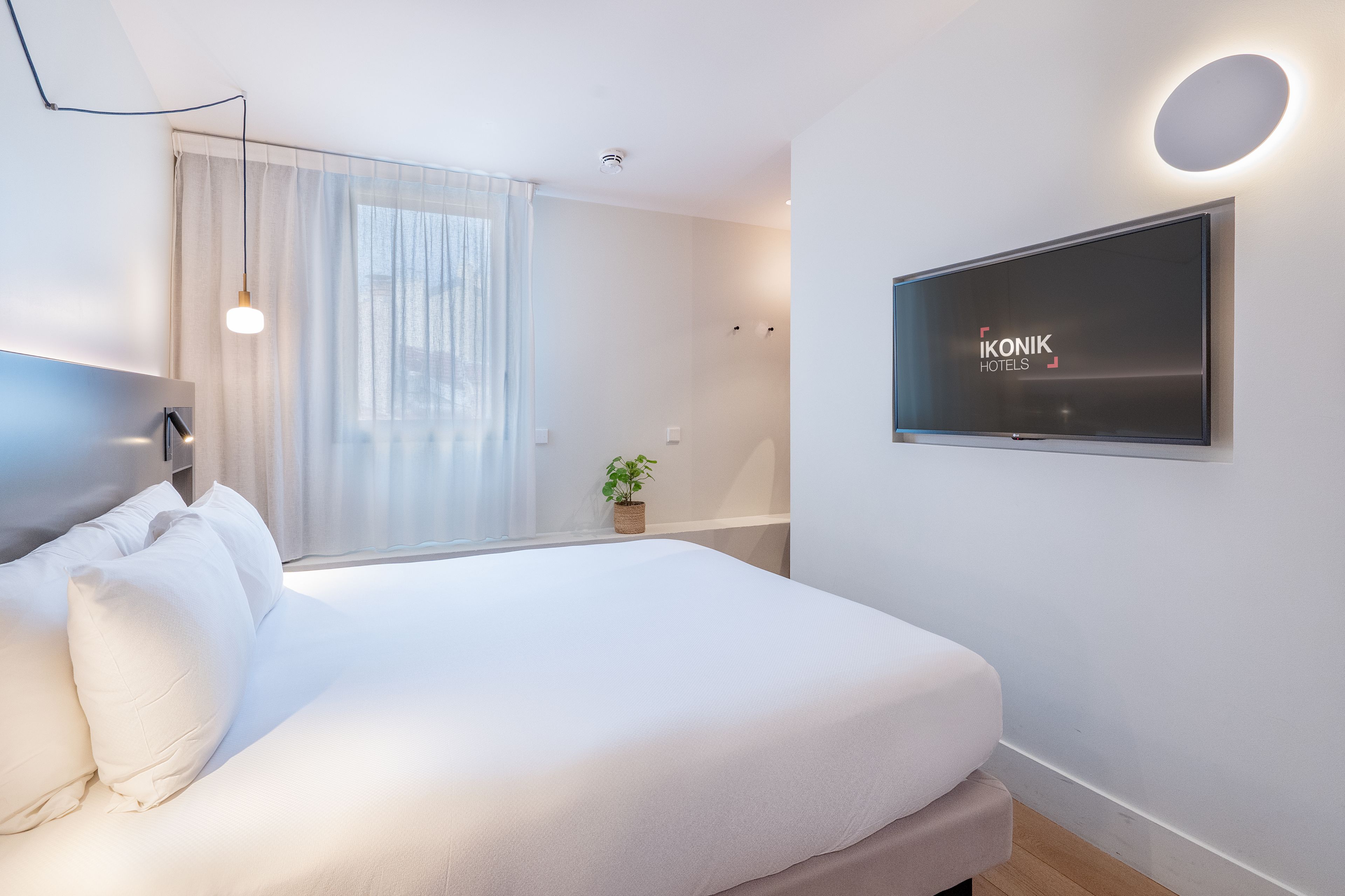Quarto de hotel moderno com uma cama grande, televisão de tela plana na parede e uma janela com cortinas finas.
