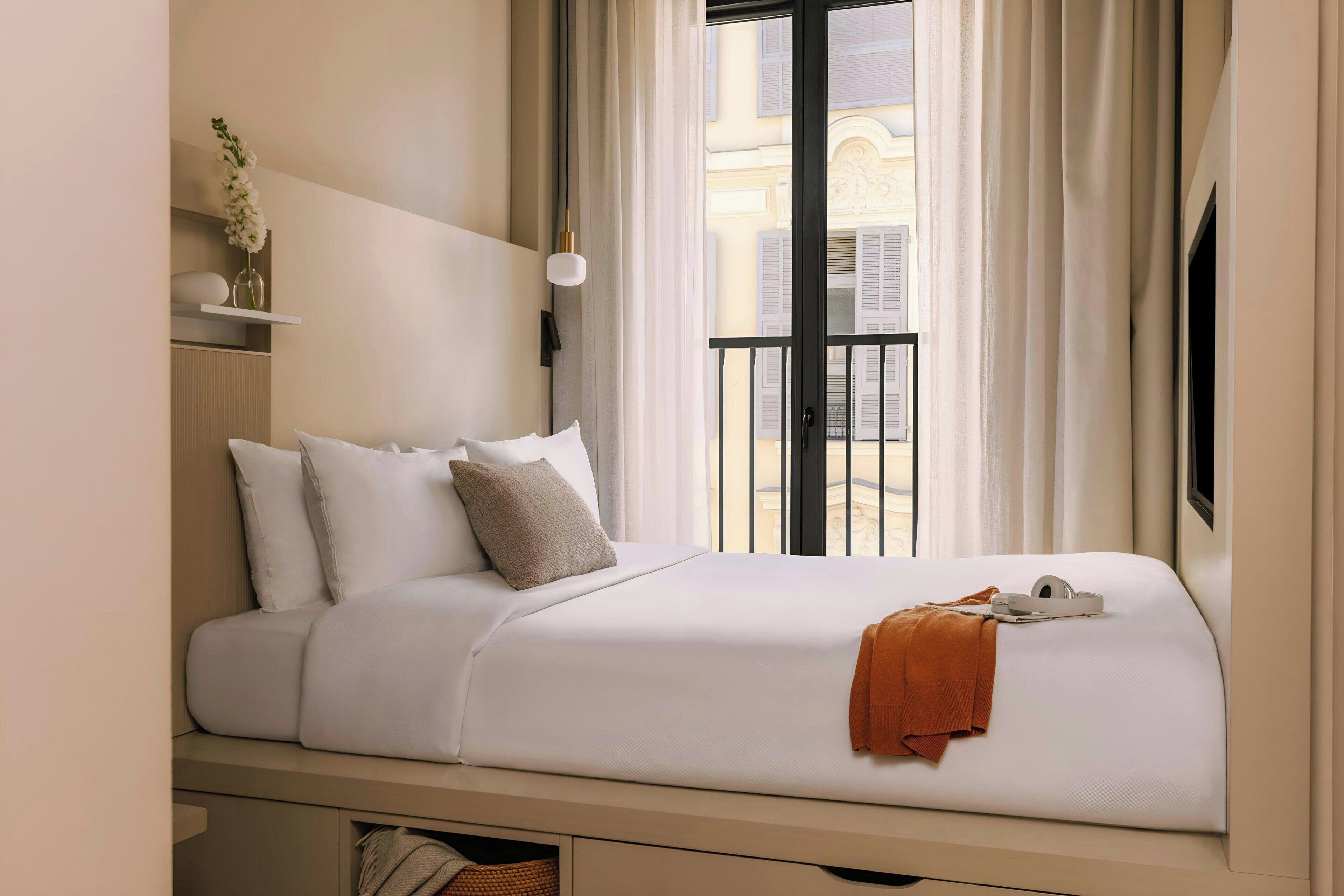Una cama bien hecha con ropa blanca y una manta marrón, un pequeño estante con un florero, y vistas a un balcón con barandilla.