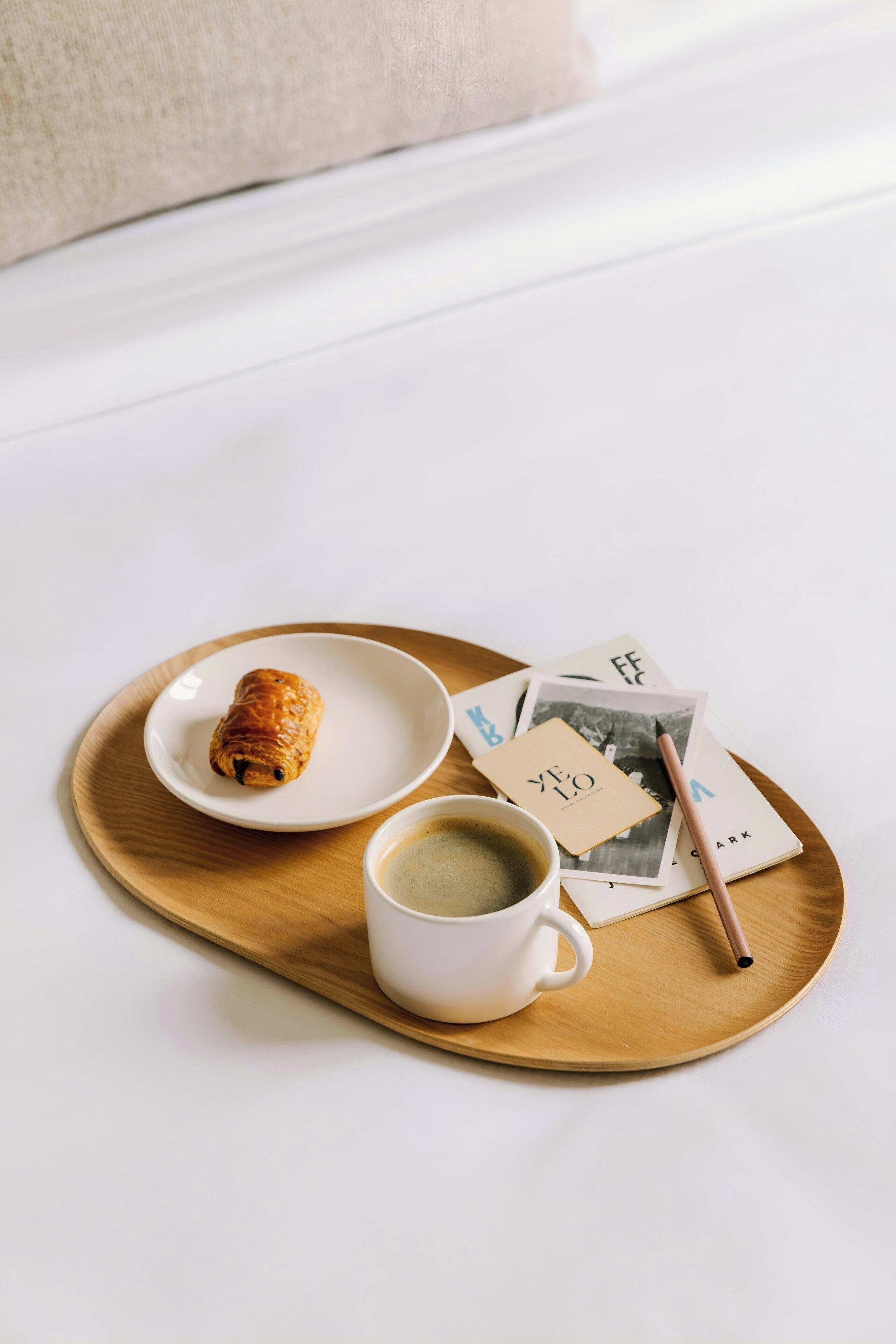 A tray with a croissant, a cup of coffee, and a book on a white table.