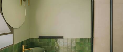 A bathroom with a green-tiled wall, a round mirror, a sink with a gold faucet, and a shower area.