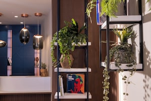 Uma área de recepção moderna com parede azul, painéis de madeira e um balcão de recepção branco. Há três luminárias pendentes, prateleiras com plantas e um quadro de Andy Warhol.