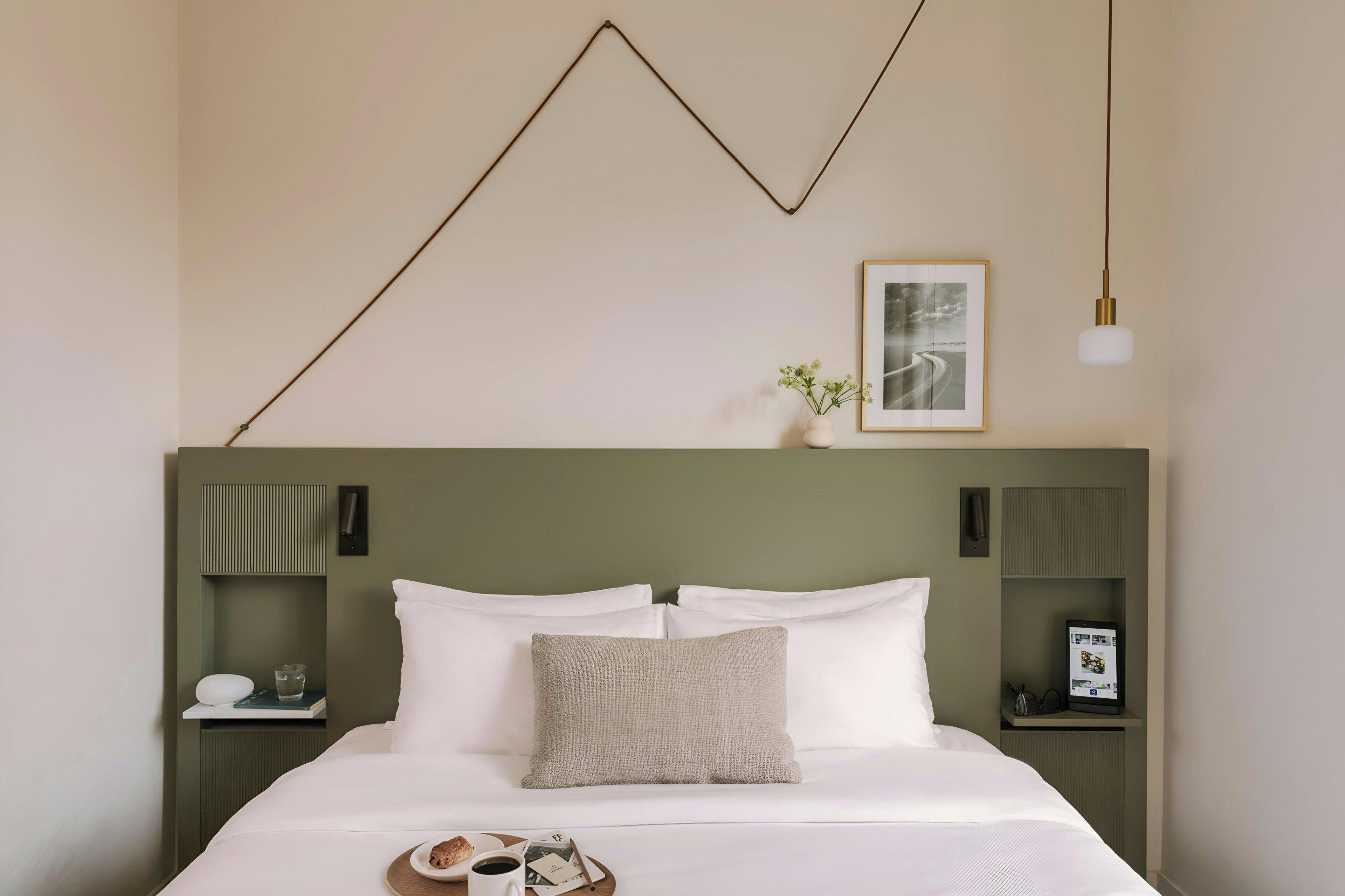 Een net opgemaakt bed met witte lakens, een grijs kussen en een dienblad met een kop en een boek. Er zijn twee nachtkastjes, elk met een kleine plant en een ingelijste foto.
