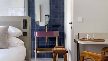 A modern hotel room with a large bed, a vanity with a purple sink, a mirror, and a wooden chair.