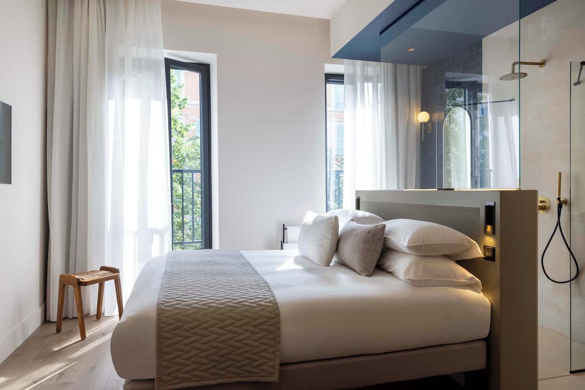 Ein modernes Hotelzimmer mit einem großen Bett, einem hölzernen Hocker, einer Duschkabine aus Glas und Blick ins Freie.