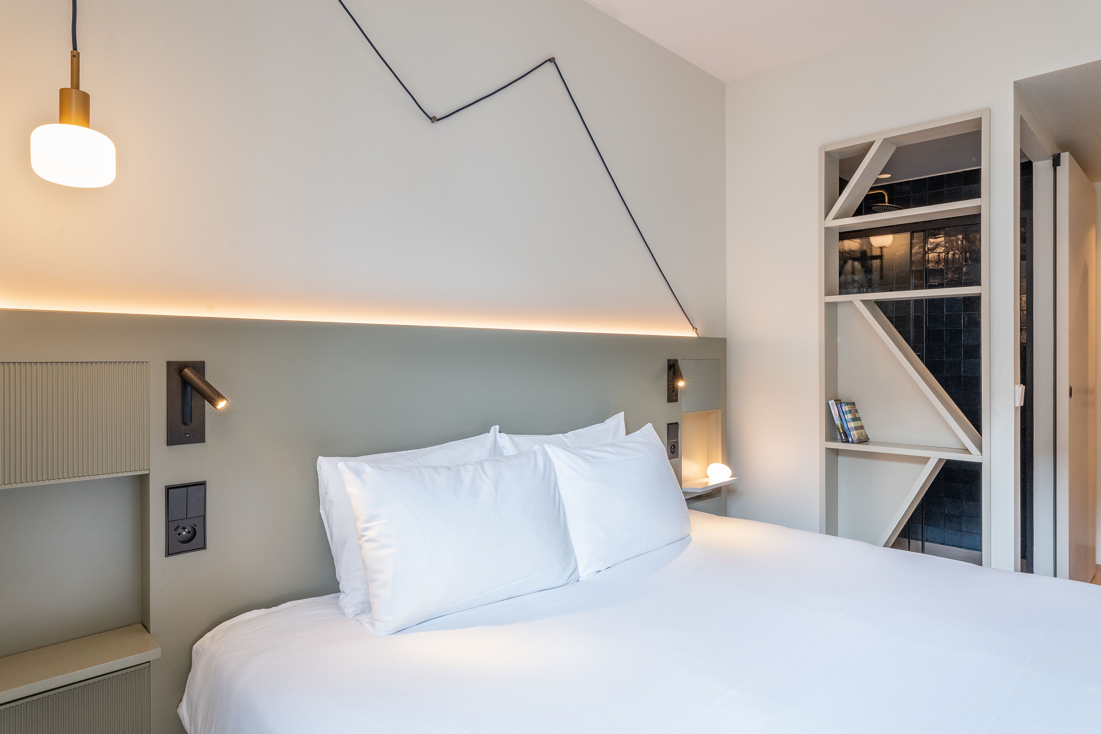 A modern hotel room with a bed, bedside lamps, a wall-mounted light fixture, and a built-in bookshelf.