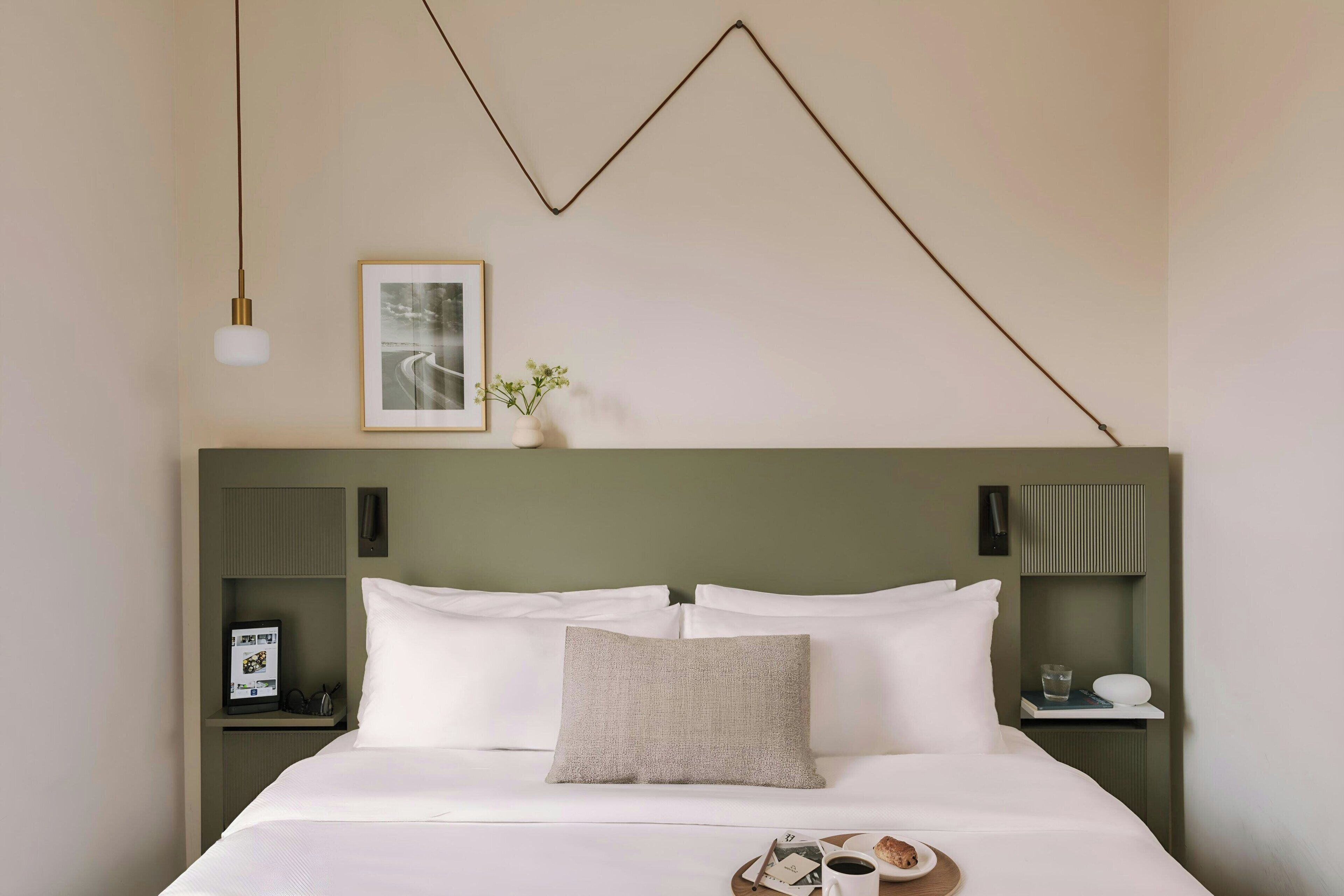 Un lit bien fait, avec du linge de lit blanc, un seul oreiller et une table de chevet sur laquelle se trouvent une tasse et un livre.