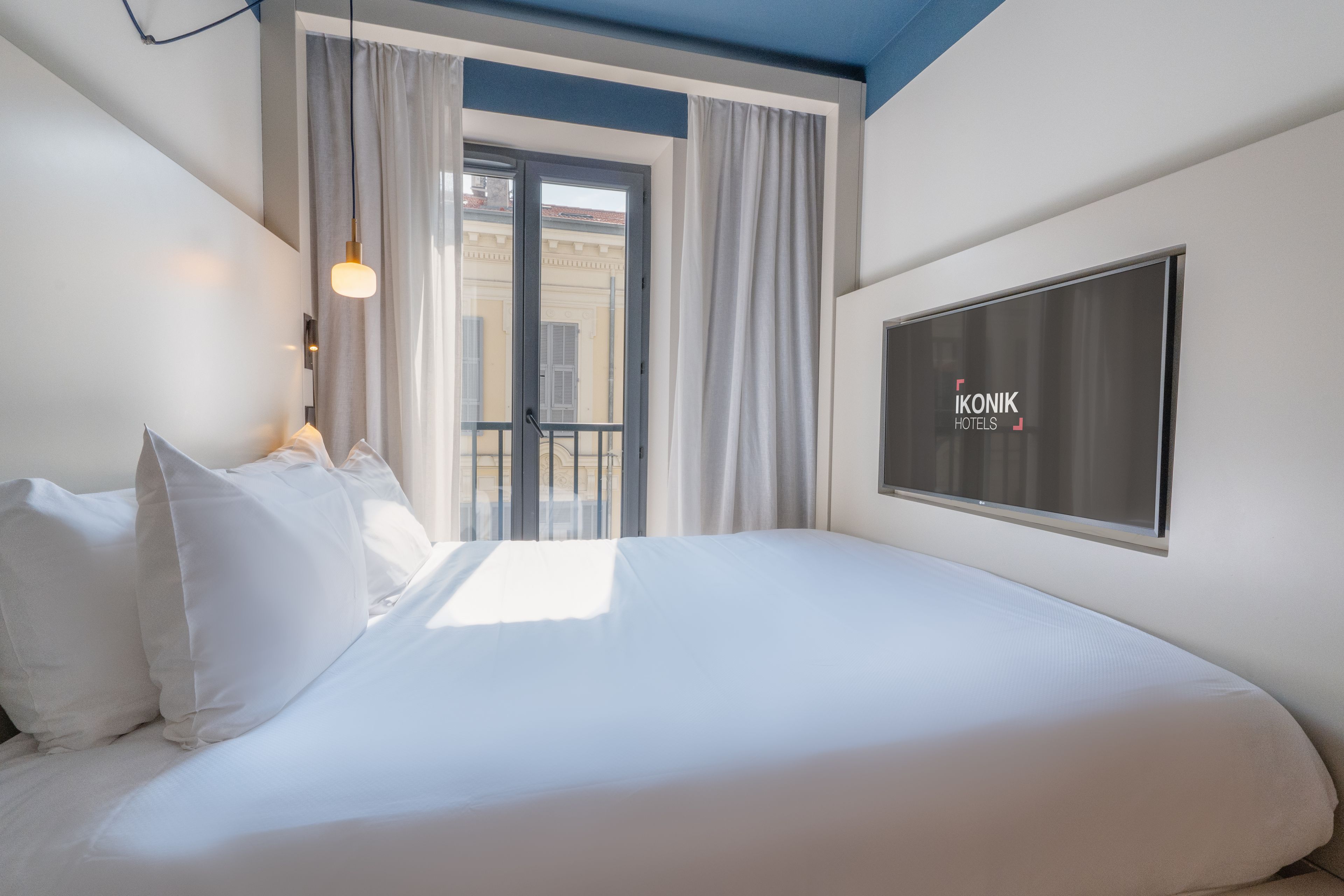 A hotel room with a bed, a TV, and a balcony with curtains.