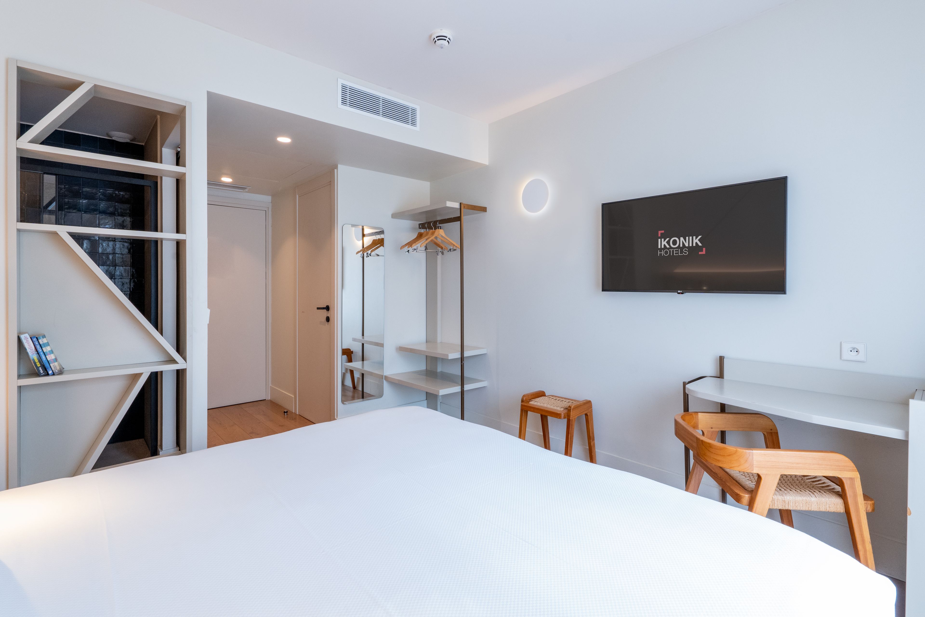 Habitación de hotel moderna con una cama grande, un televisor montado en la pared, un armario con estantes y una pequeña zona de estar con una silla y una mesa.
