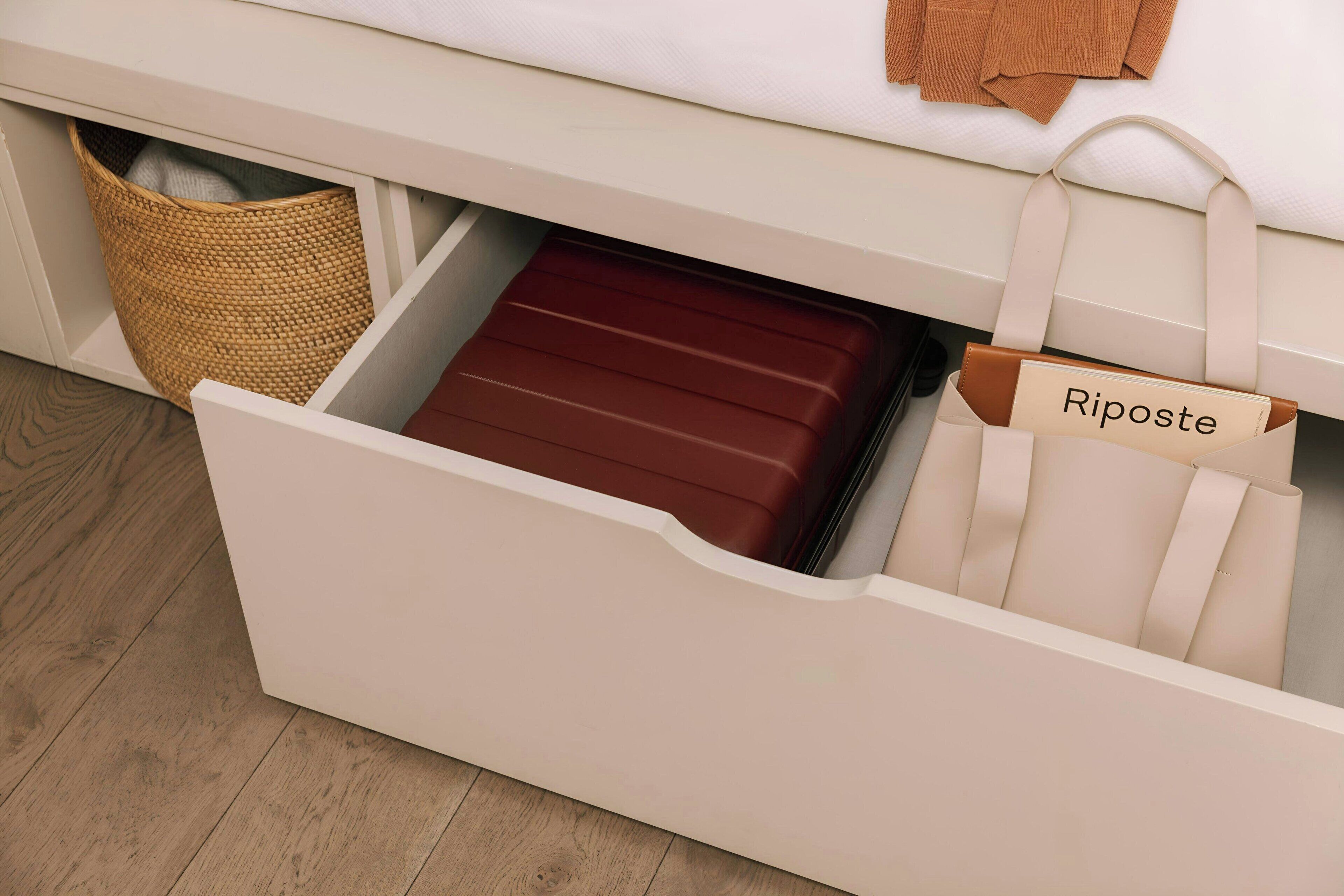 Un cajón blanco con un cesto de mimbre y una maleta marrón con la etiqueta 'Riposte' en su interior.