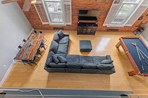 A room with a wooden pool table, a sectional sofa, a dining table with chairs, and a television against a brick wall.