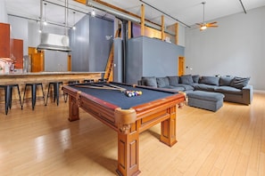 A pool table with a wooden finish, a bar area with stools, and a gray sofa in a modern living space.