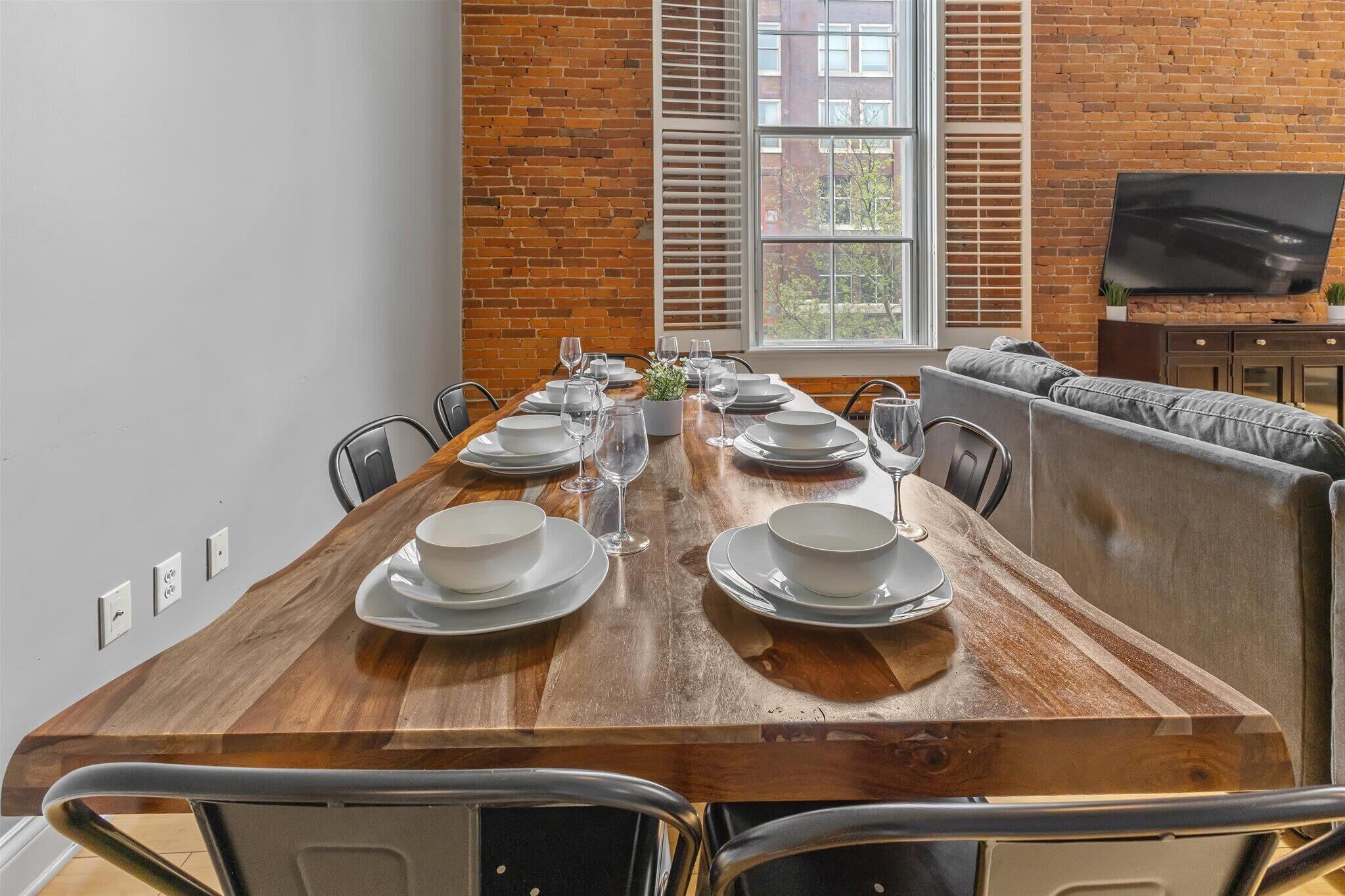 A dining area with a long wooden table set for six, flanked by a sofa and a television.