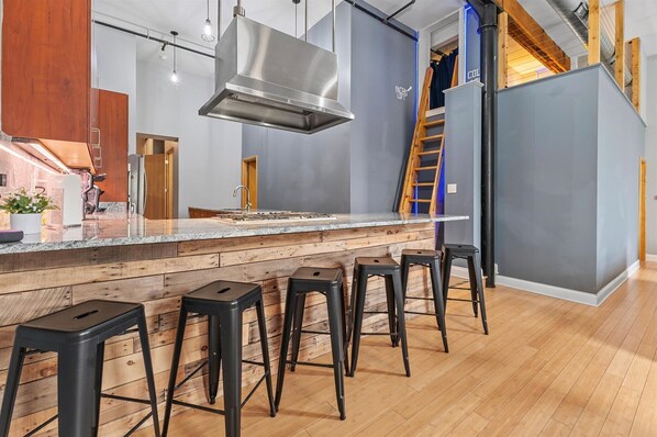 A modern kitchen with a central island, bar stools, a stainless steel range hood, and a wooden staircase.