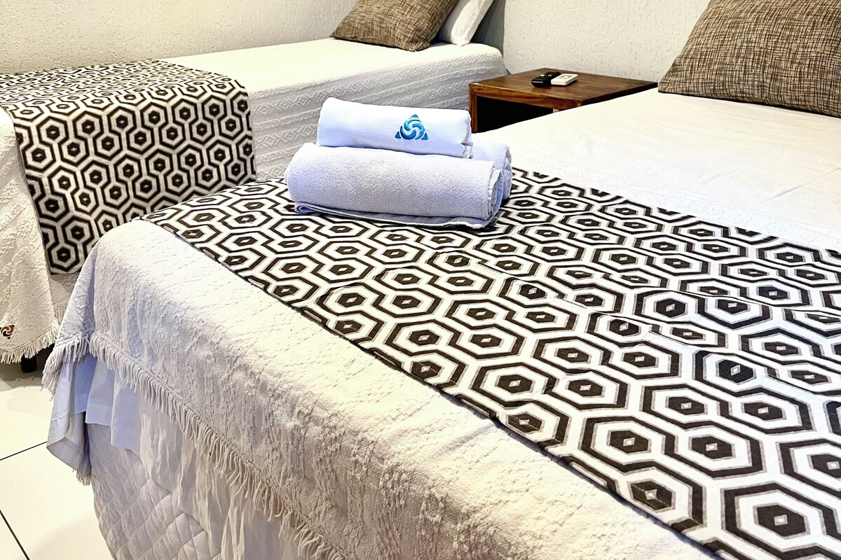 Habitación de hotel con cama, dos almohadas estampadas, una toalla doblada, una mesita de noche y un aire acondicionado instalado en la pared.