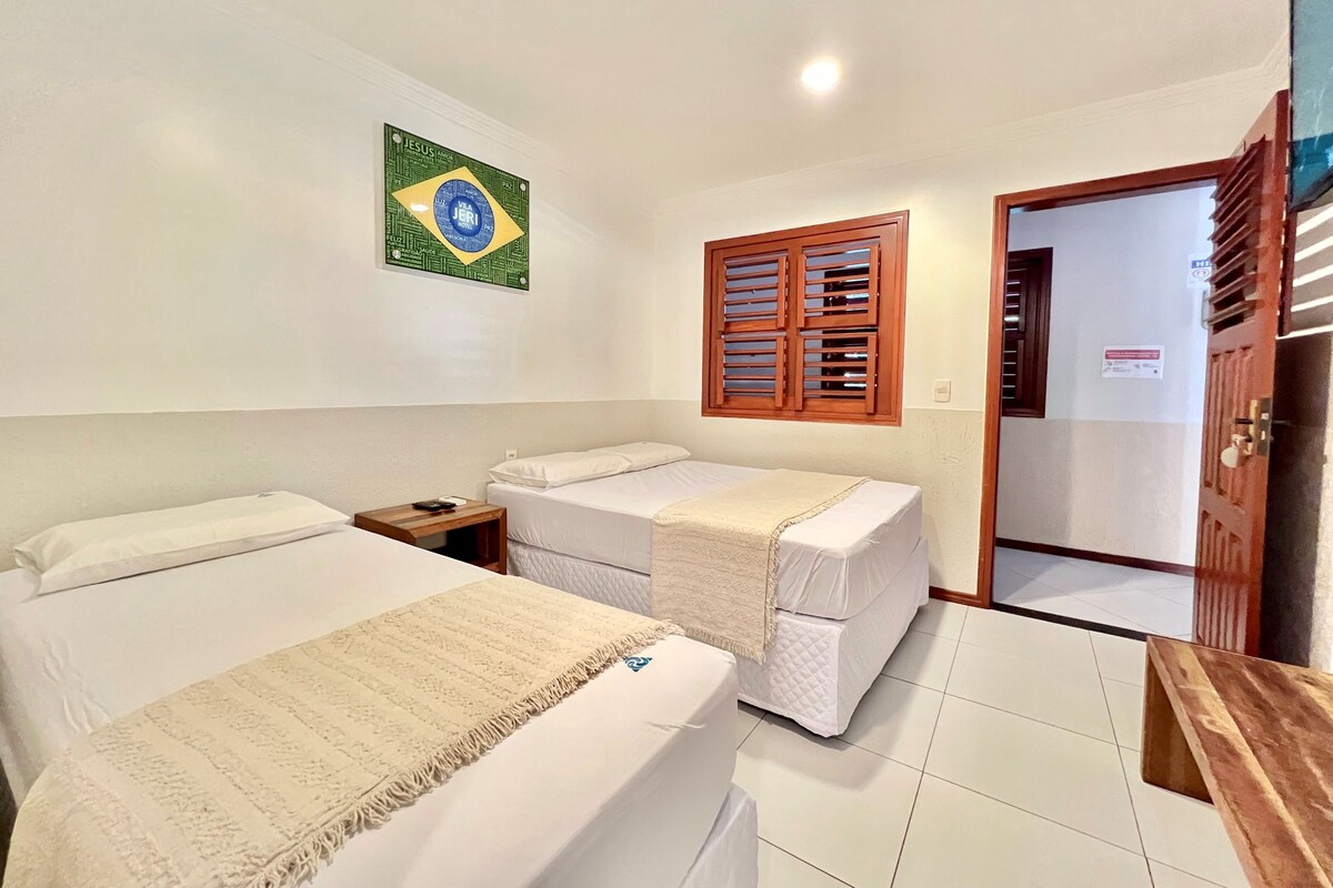 Habitación de hotel con dos camas, una puerta de madera, una ventana con contraventanas y un letrero en la pared con una bandera.