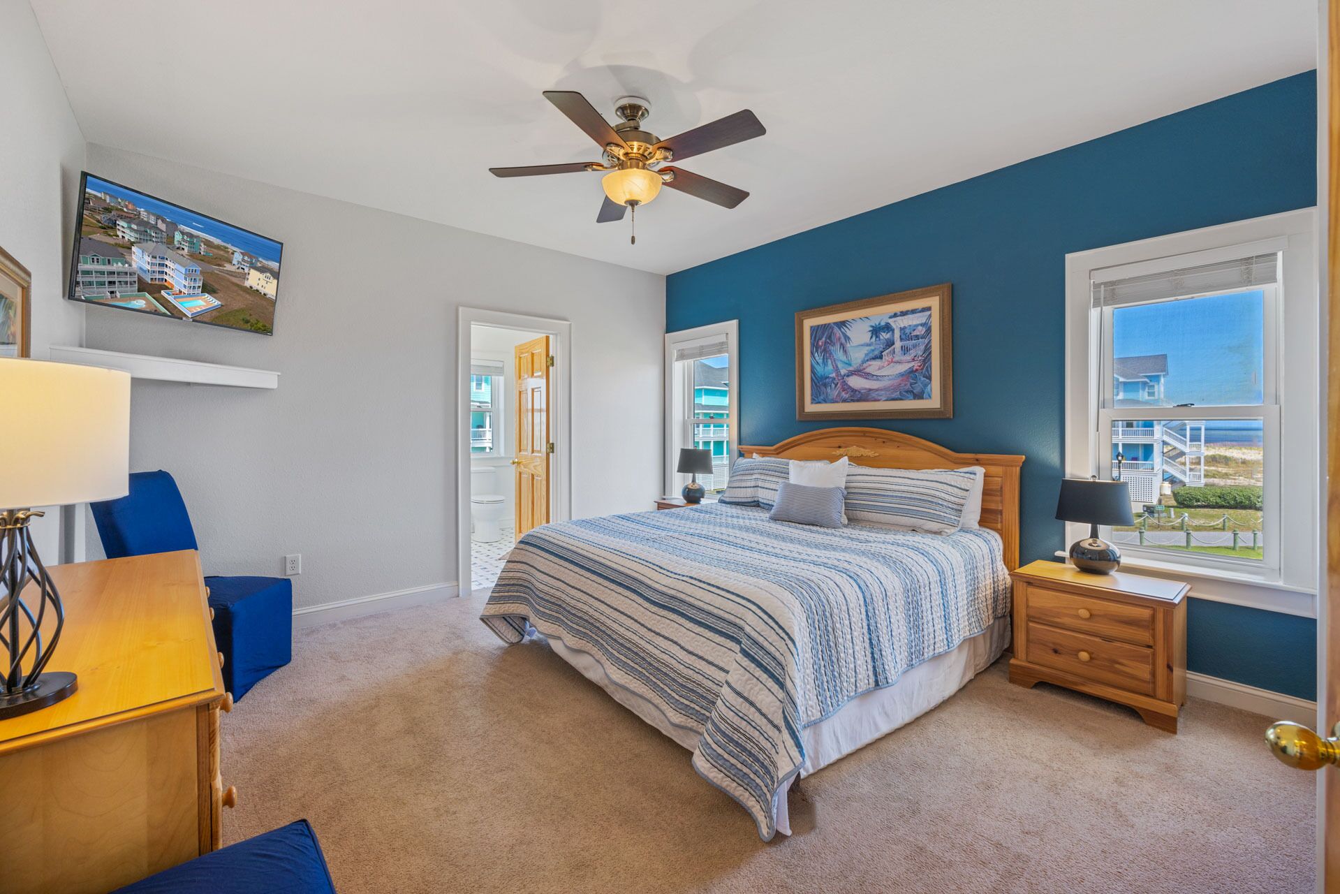 A bedroom with a bed, a ceiling fan, a window with a view, a painting, and a door leading to another room.