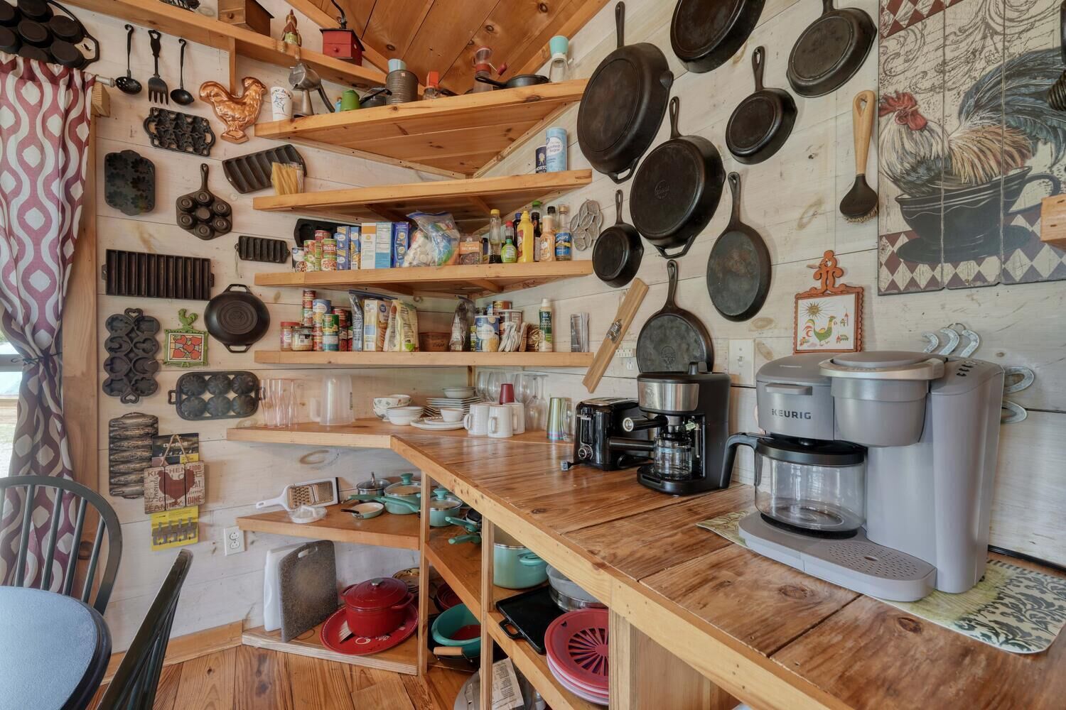 Una cocina con estantes de madera que contienen diversos utensilios, ollas y sartenes. Hay una cafetera y un hervidor en la encimera.