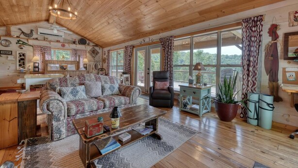 Ein gemütliches Blockhausschlafzimmer mit Holzbalkendecke, einem gemusterten Sofa, einem schwarzen Ledersessel, einem kleinen Tisch und einem großen Fenster mit Blick ins Freie.