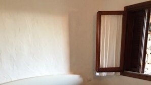 A bed with a blue bedspread and pillows, a nightstand with a lamp, and a window with curtains. - Bilocale Vista Mare (San Teodoro)
