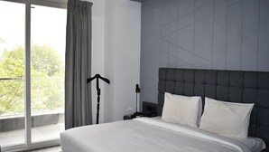 A modern hotel room with a large bed, a nightstand, a lamp, and a view of greenery through the window. - Bedchambers Serviced Apartments, MG ROAD (Gurugram)