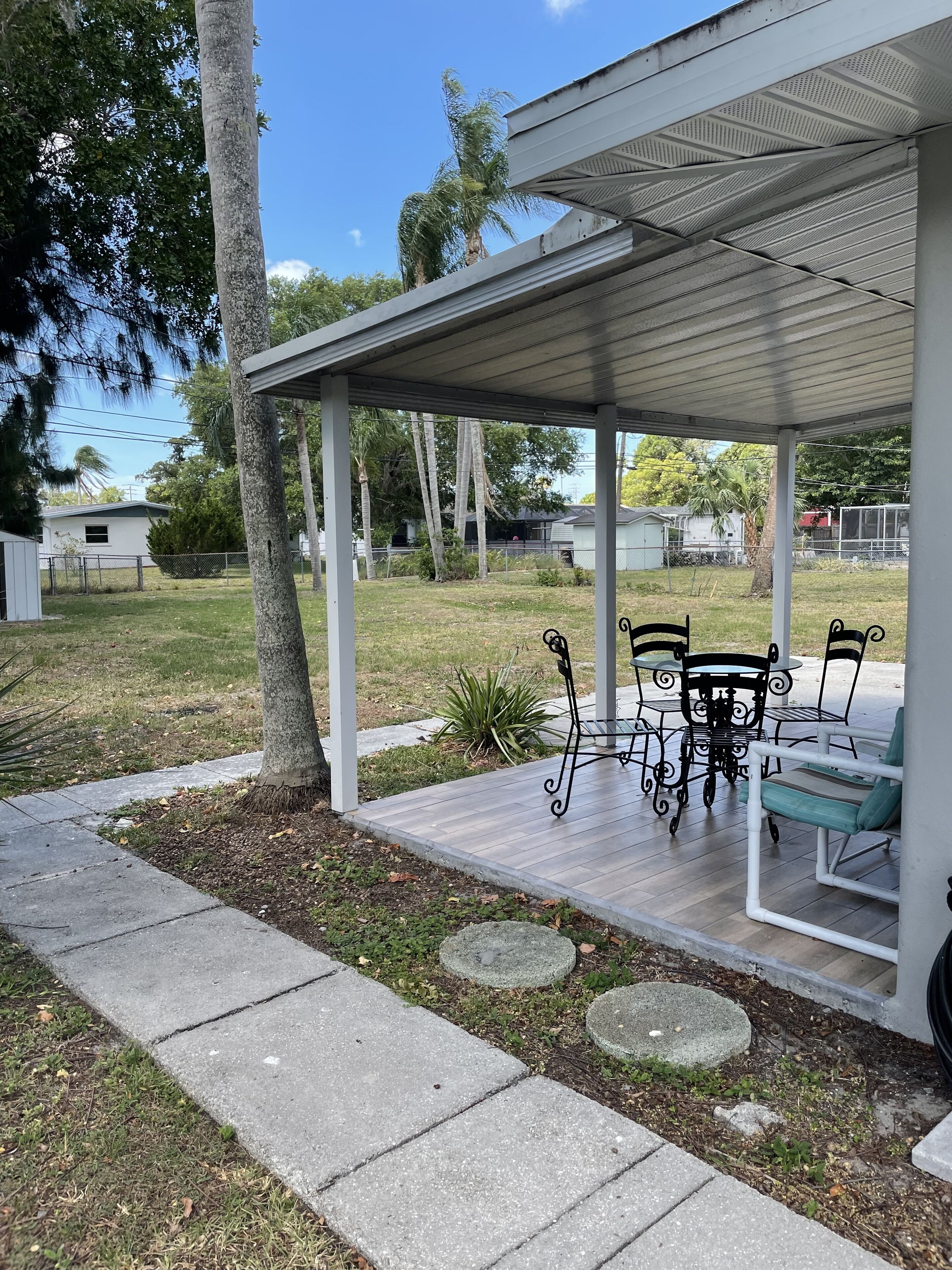 A covered patio with a table and chairs, a stone pathway, and a view of a residential area with trees and a house.