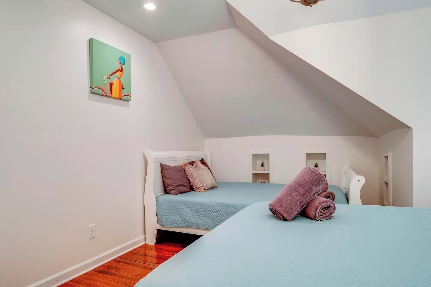 A small, modern bedroom with a sloped ceiling, a bed with a blue bedspread, a white headboard, and a painting on the wall.