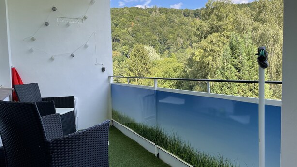 A balcony with a view of a forested hillside, a white railing, and a table with chairs.