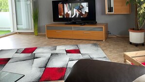 A modern living room with a television, a wooden cabinet, a potted plant, and a large window.