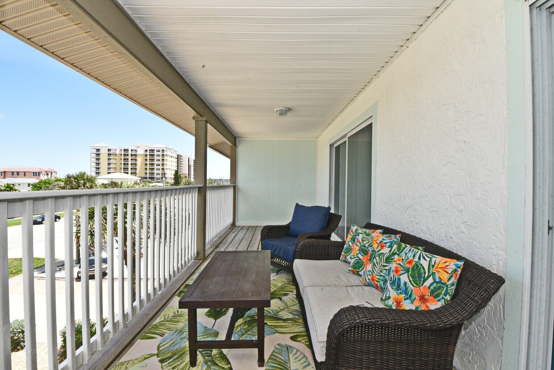 A balcony with a wicker sofa, a small table, and a view of buildings in the background.
