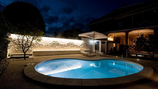 A swimming pool with a stone wall and lounge chairs, surrounded by a building with a covered patio. - VILLA IARA IN CERVETERI WITH PRIVATE SWIMMING POOL (Cerveteri)