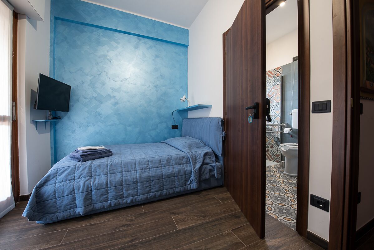 Une chambre avec un mur bleu à texture, un lit recouvert d’une couette bleue, un téléviseur fixé au mur et une porte en bois donnant sur une salle de bain.