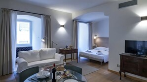 Ein Hotelzimmer mit Essbereich, einer Couch, einem Bett, einem Fernseher und einem Fenster mit Vorhängen.