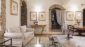 A cozy living room with a white sofa, a glass coffee table, and a laptop. There are framed pictures on the walls, a stone wall, and a window with curtains.