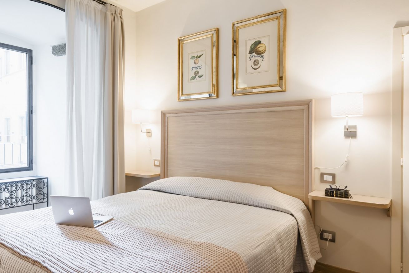 Una camera d'albergo con un letto, un portatile sul letto, due quadri appesi alla parete, un comodino con una lampada e una finestra con tende.