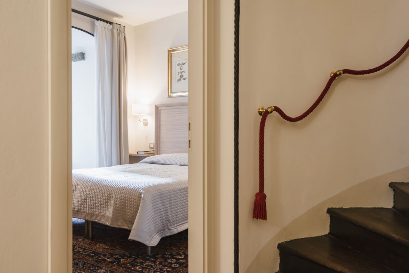 A hotel room with a bed, a nightstand, a lamp, a framed picture on the wall, and a red tassel on the staircase.