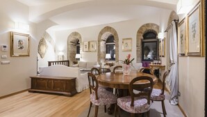 A spacious living room with a wooden dining table, floral chairs, and a sofa. There are arched doorways and framed artwork on the walls.