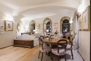 Ein geräumiges Wohnzimmer mit einem Holzes Esstisch, floralen Stühlen und einer Couch. Es gibt gewölbte Durchgänge und gerahmte Kunstwerke an den Wänden.