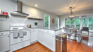 A modern kitchen with white cabinets, a stainless steel stove, and a dining area with a table and chairs. - Treetop house with direct access to bike trails and small walk to Lake Avalon (Bella Vista)